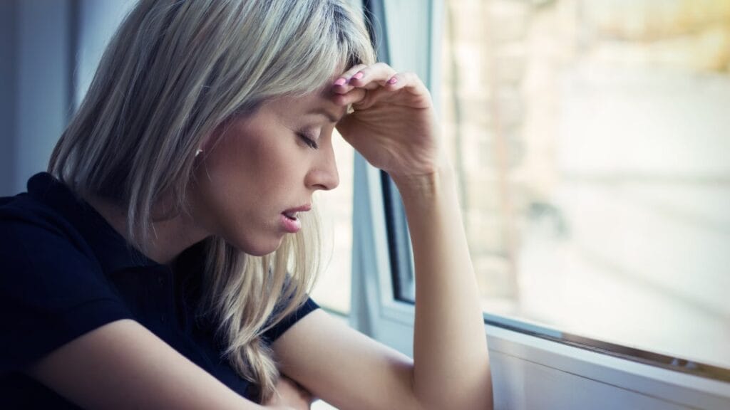 Femme pensive près d’une fenêtre, main au front — remise en question de ses choix amoureux.